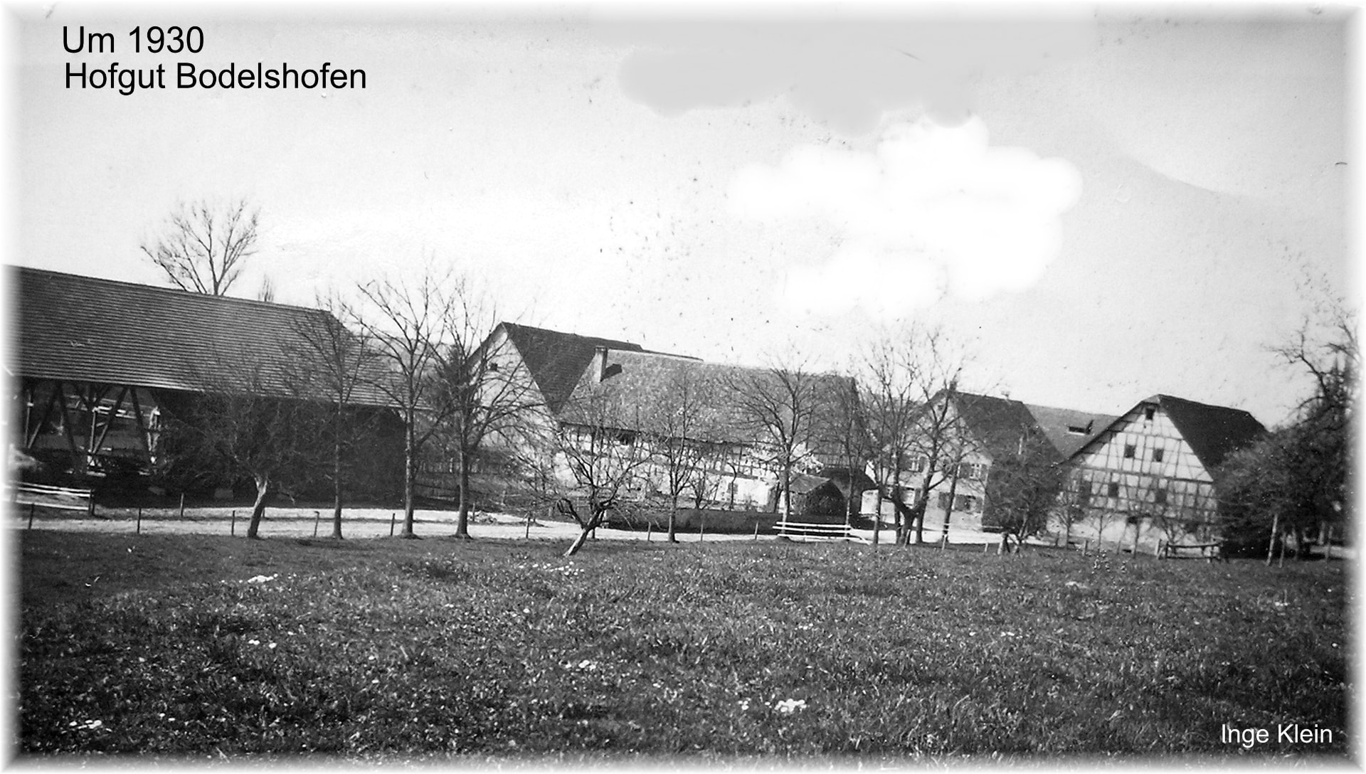 Historische Schwarzweißaufnahme der Hofgebäude mit Fachwerk und Scheune hinter einer Wiese mit kahlen Bäumen und einfachem Zaun