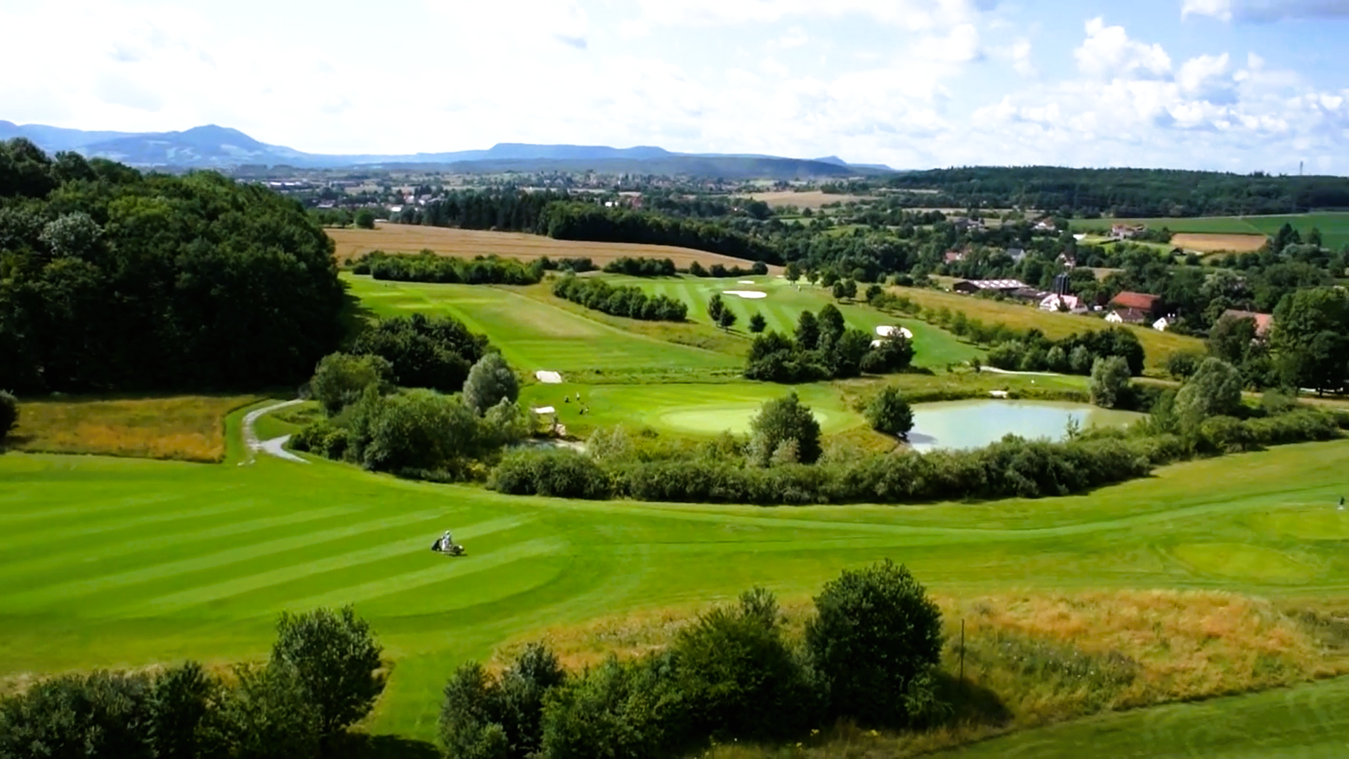 Luftbild des Golfplatzes mit Fairways, Sandbunkern, Teich und bewaldeten Hügeln im Hintergrund
