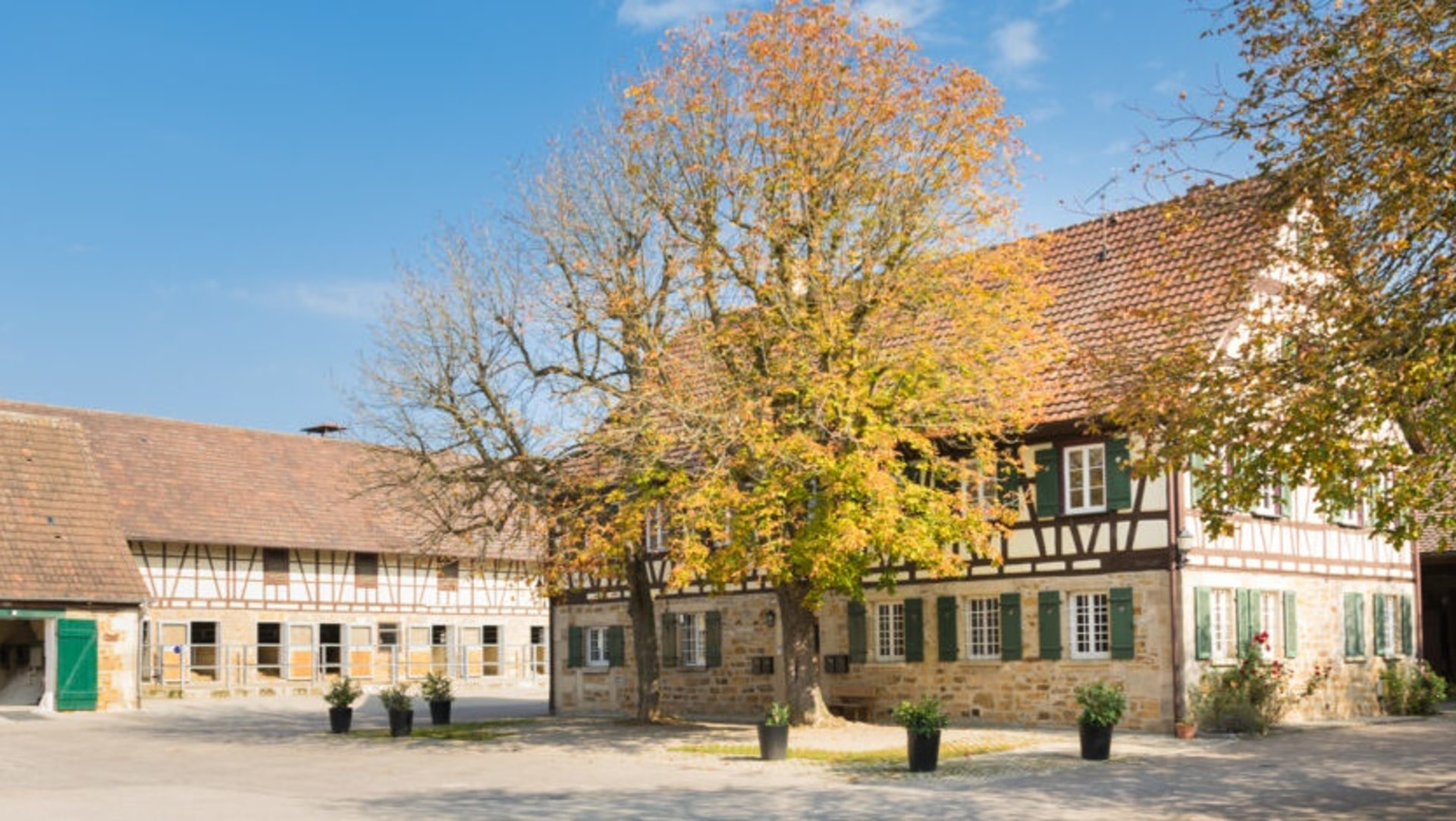 Gepflasterter Hof mit Fachwerkhaus, grünen Fensterläden und Stallgebäuden im Herbstlaub