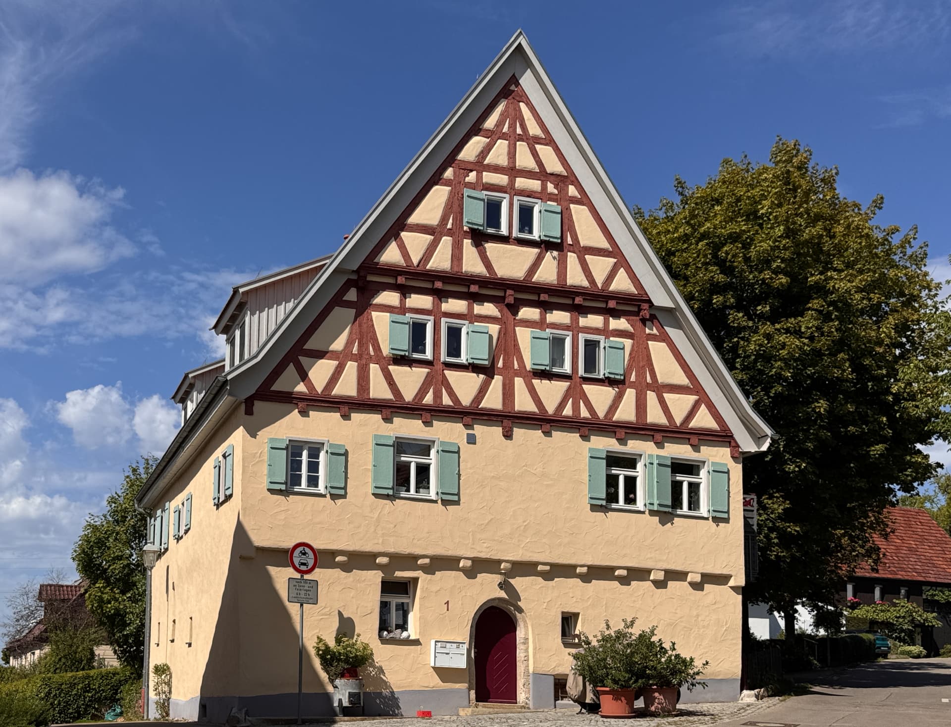 Fachwerkhaus mit steilem Satteldach, Gaube und grünen Klappläden vor gepflastertem Vorplatz im Sommer