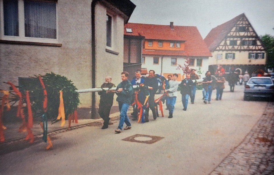 Farbfoto einer Gruppe von Männern und Jugendlichen, die einen geschmückten Maibaum mit bunten Bändern eine Dorfstraße entlangtragen