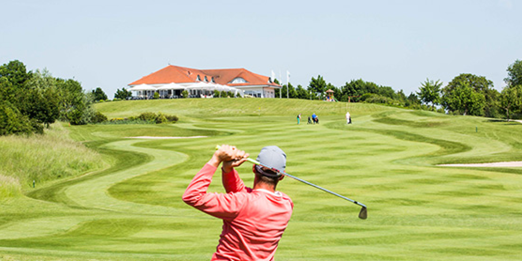 Golfspieler beim Abschlag auf dem Fairway, im Hintergrund das Clubhaus auf der Anhöhe