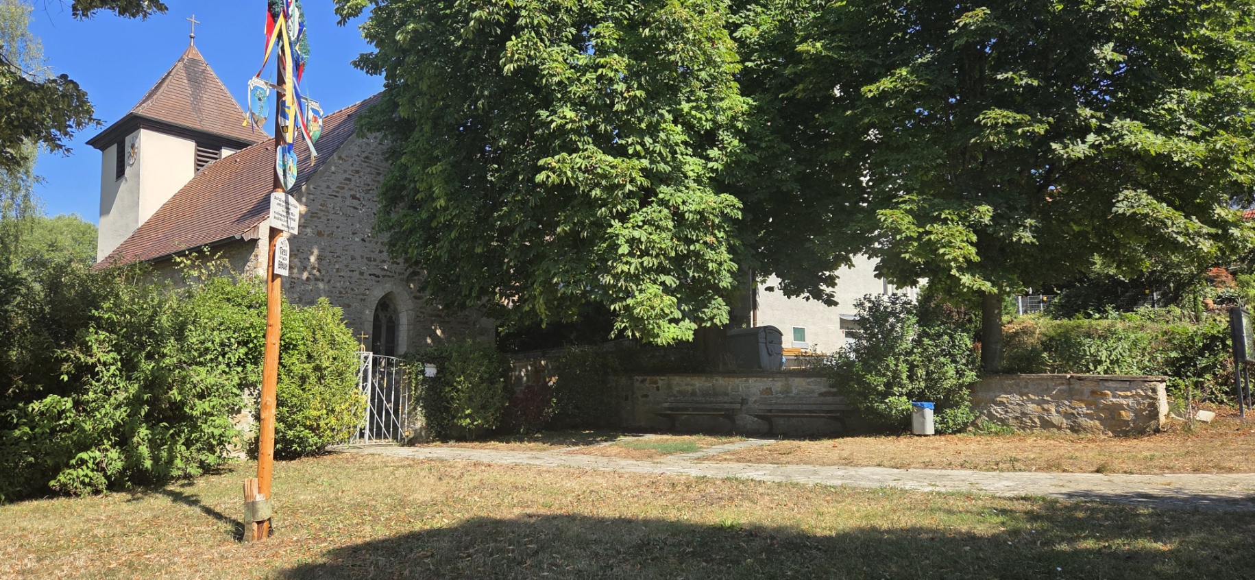 Jakobskirche aus Bruchstein mit Turm und Maibaum auf dem Kirchplatz mit Bänken und Bäumen im Sommer