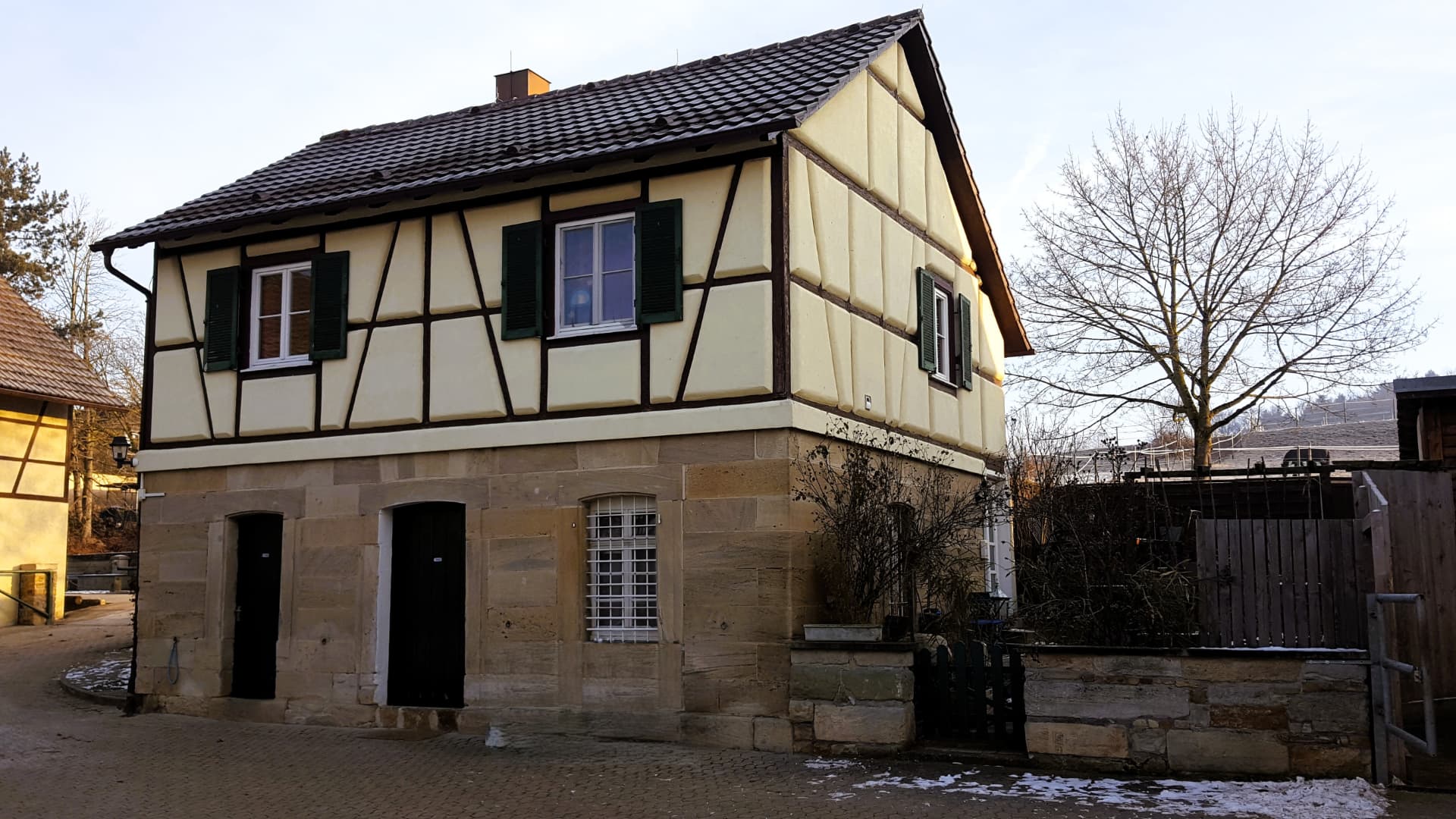 Fachwerkhaus mit Sandsteinsockel an einem gepflasterten Hof im Winter, mit Schneeresten und kahlen Bäumen