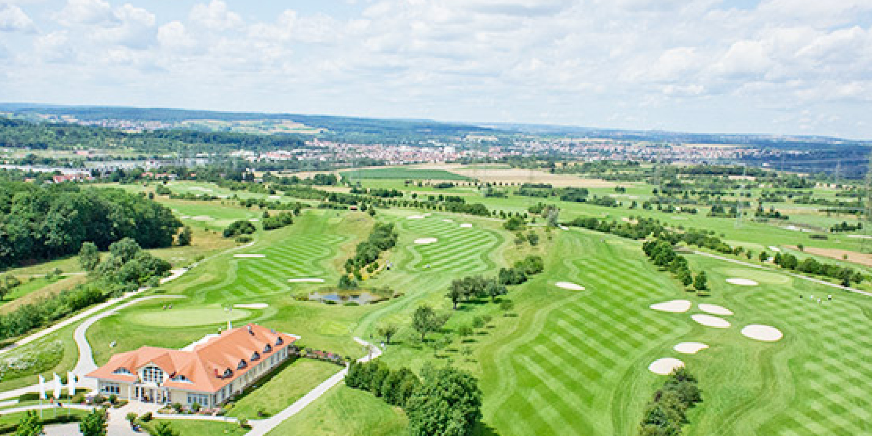 Luftbild des Golfplatzes mit Teich, Sandbunkern und Clubhaus im Vordergrund, dahinter weite Landschaft